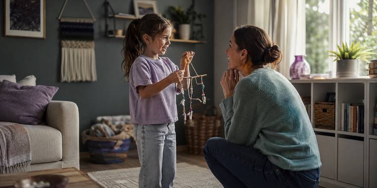 A girl holding up a craft she made to show her mother, who is crouching to look